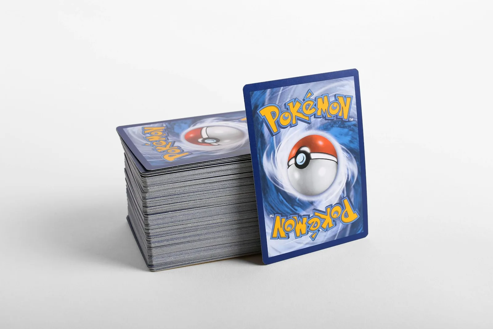 A tall stack of Pokemon cards face down on a white surface with one card standing upright against the side of the stack