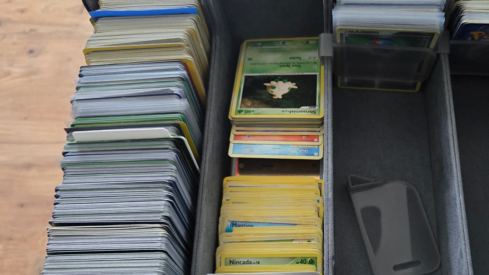Rows of Pokemon cards inside a gray storage box on a wood table: a packed row of several hundred cards stood on edge at the left, small face up stacks part way through sorting in the middle, and toploaders and a Trainer card in the row behind